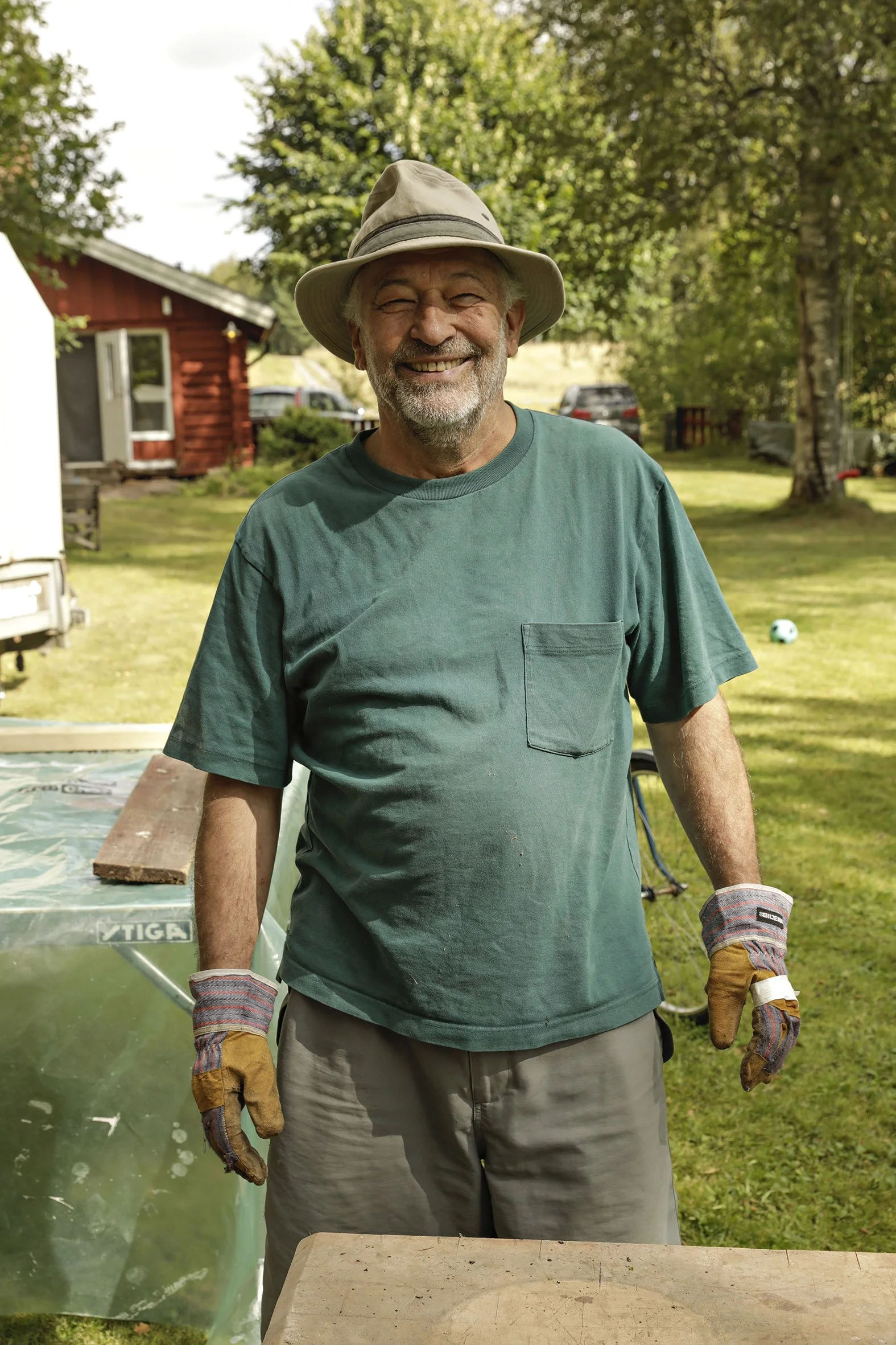 Abbi Patrix smiling outdoors, wearing a wide-brimmed hat, green t-shirt, and work gloves. There are trees, a red house, and cars in the background.
