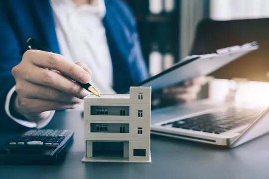 Person in business attire holding a pen over a miniature model of a building on a desk with a calculator, laptop, and documents.