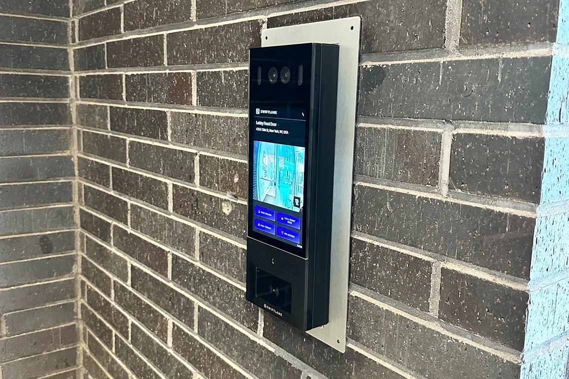 An intercom system with a touchscreen display mounted on a brick wall, showing a camera feed and options for access control.