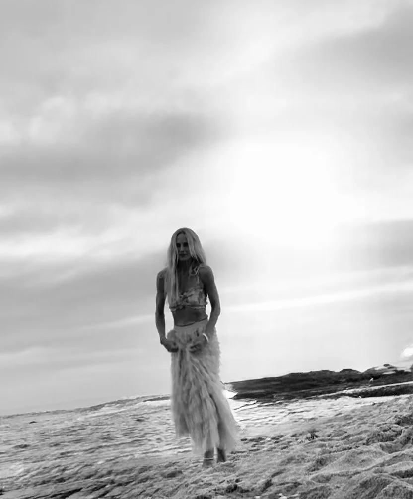 A woman with long, wavy hair stands on a sandy beach near the water, wearing a skirt and crop top, under a cloudy sky in black and white.
