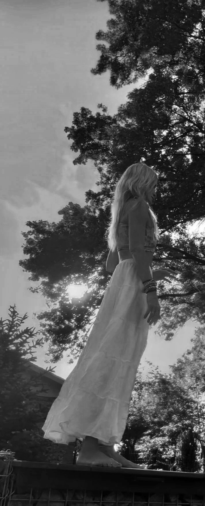 A jessi with long, wavy hair wearing a flowing skirt and bracelets, standing outdoors on a platform with trees and a cloudy sky in the background nature grounds.  manifest relationships through grounding yourself