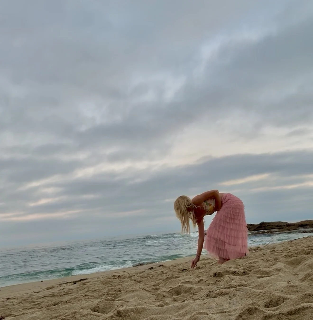 Jessi in a pink tiered skirt and patterned top bending down on a sandy beach near the ocean with cloudy sky overhead.
