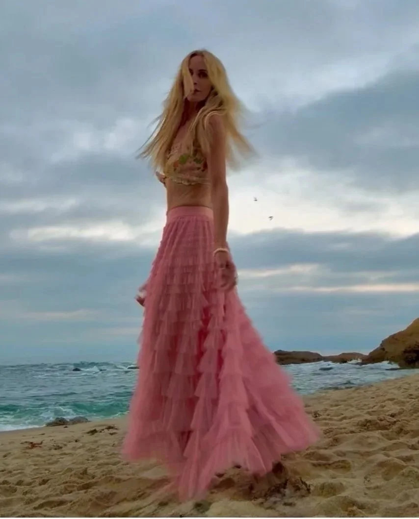 A woman with long blonde hair standing on a beach, wearing a pink full-length ruffled skirt and a sleeveless top.