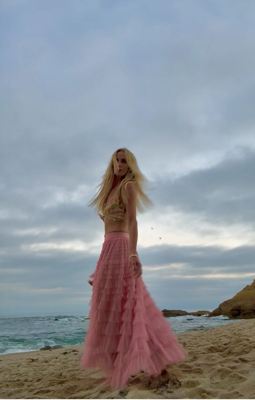 Jessi Jaksha with long blonde hair wearing a pink layered skirt and a floral crop top standing on a sandy beach near the ocean with a cloudy sky overhead.  in the clouds there is a force and frequency that always faith and believe in surrender