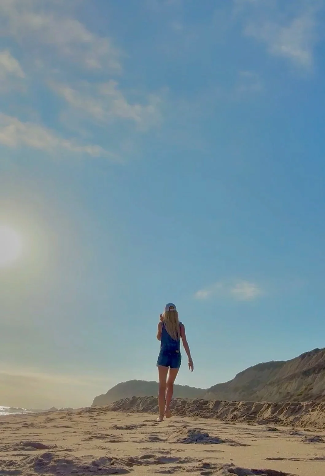 Jessi Jaksha walking on a sandy beach during sunrise or sunset with the sun low on the horizon and hills in the background. all just walking home.  don't look back, be