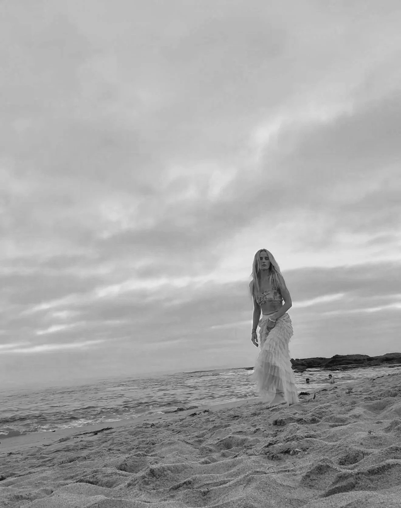 A woman in a long skirt and crop top walking on a sandy beach near the ocean, with cloudy sky overhead.