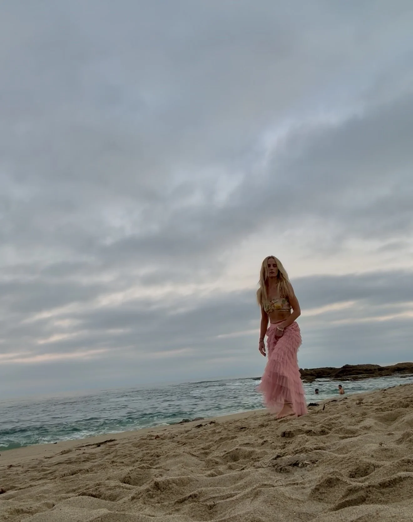 A woman in a pink ruffled skirt and a yellow patterned top standing on a sandy beach with an ocean and cloudy sky in the background.