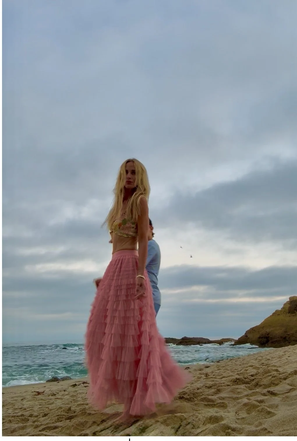 Jessi Jaksha with long blonde hair wearing a crop top and a long pink layered skirt standing on a sandy beach with the ocean and cloudy sky in the background. receiving what is possible