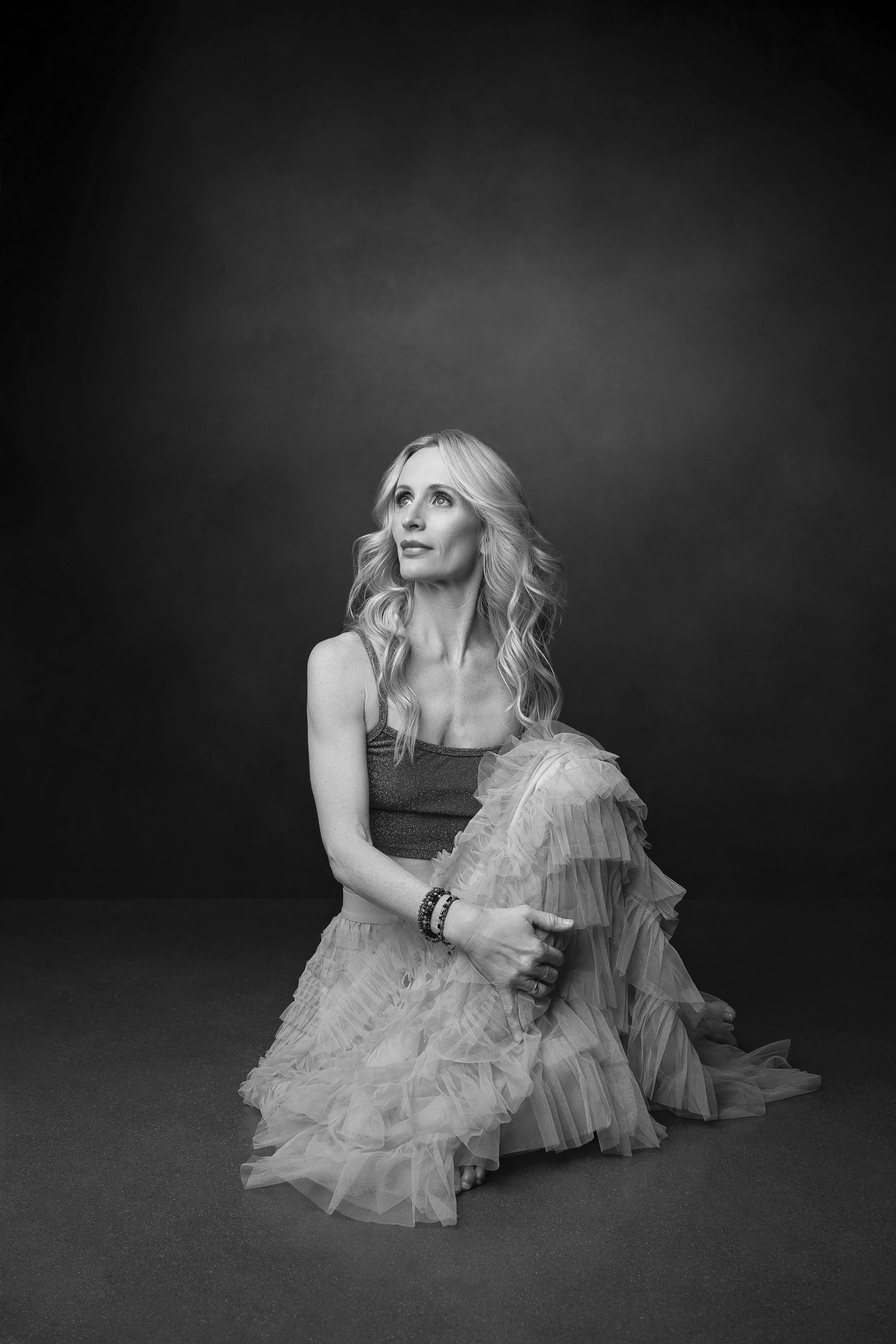 Black and white photo of a woman with long, wavy hair, sitting on the floor, holding her skirt, looking up and to the side, against a dark background.