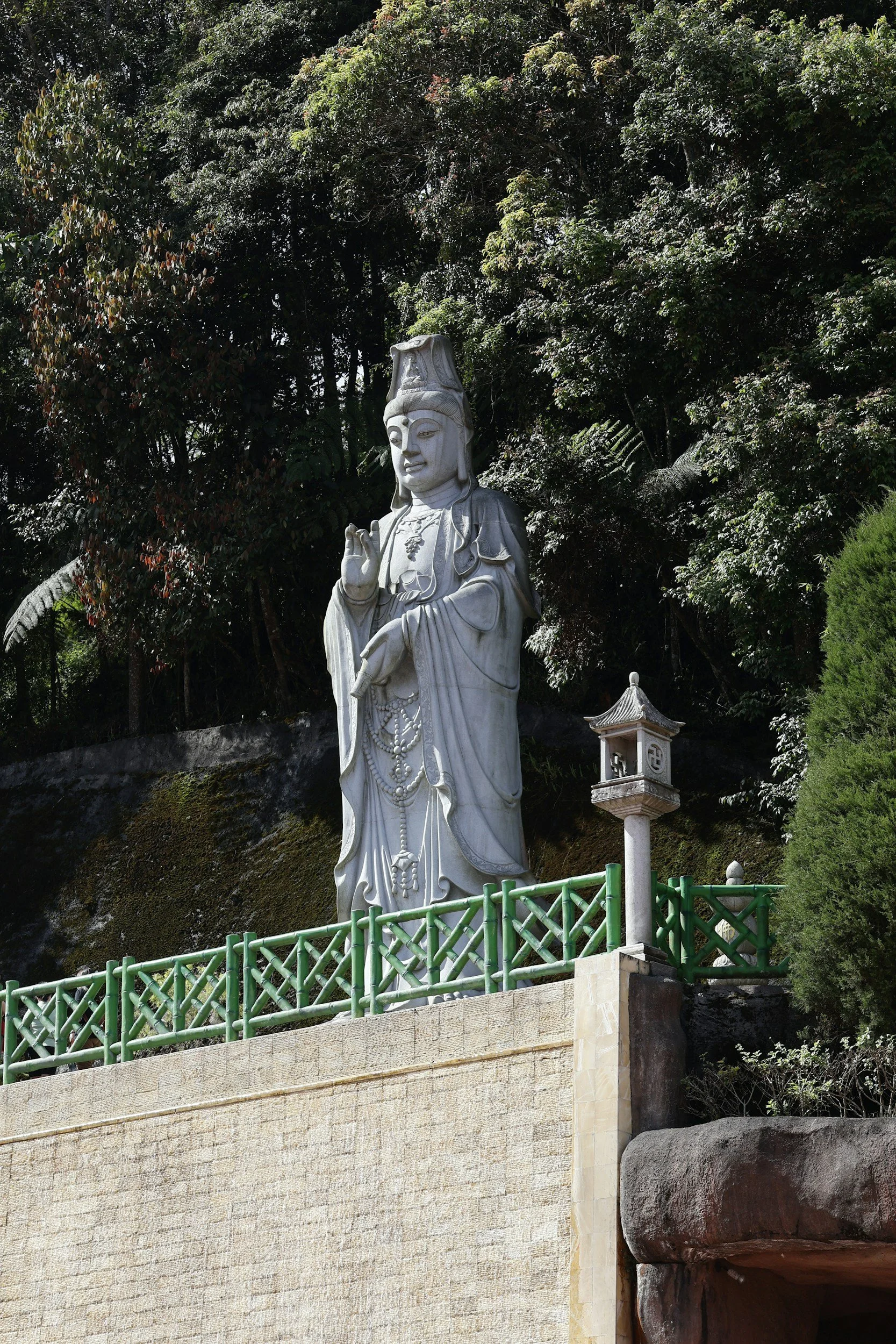 A large white statue of a serene Asian figure, likely a deity, positioned outdoors against dense green trees. The statue features an ornate headdress and traditional robes, with one hand raised in a gesturing or blessing position. A small stone lantern is present nearby, and a green fence surrounds the statue on a stone platform.