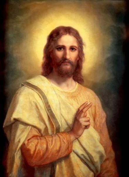 A painting of Jesus Christ with shoulder-length hair, a beard, and a warm expression, wearing a yellow and orange robe, with a glowing halo behind his head, making a blessing gesture with his right hand.