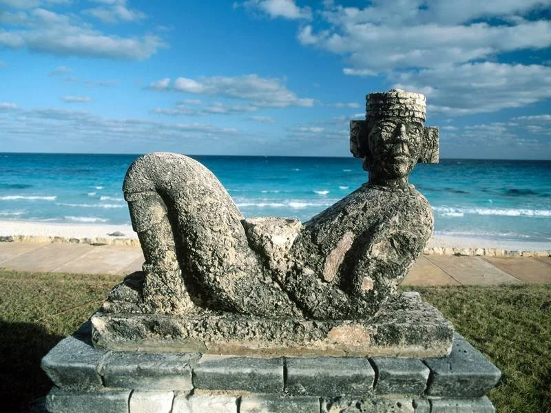 Stone sculpture of a reclining figure with a carved face and headdress, set against a beach and ocean background.