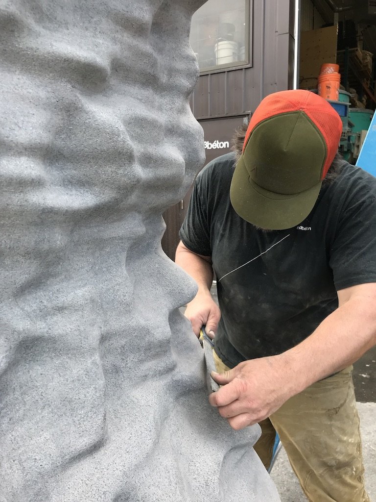 Un homme avec un casque rouge et vert sculpte une statue en pierre ou en béton, vue de profil.
