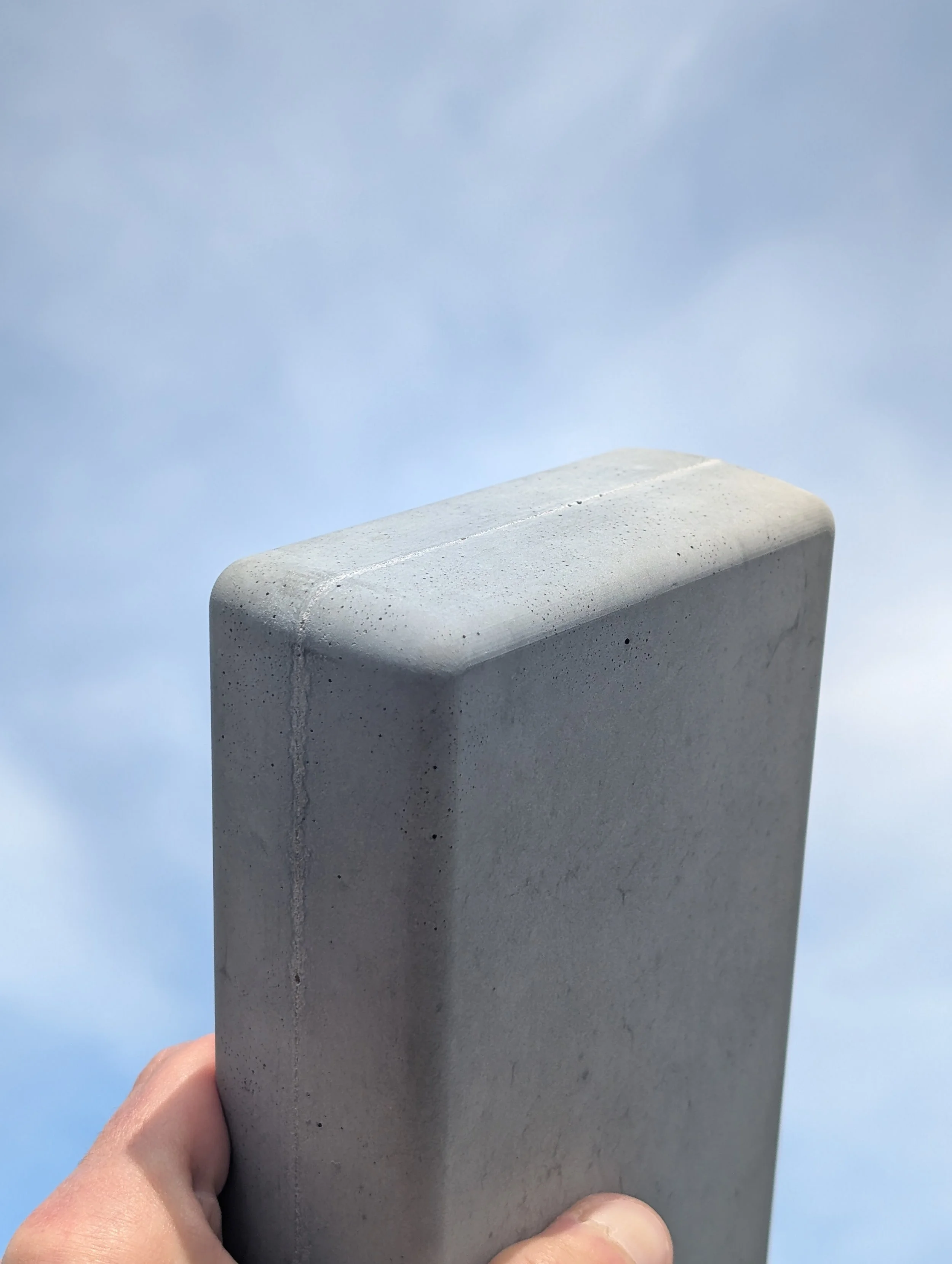Une main tenant une grande dalle de béton contre un ciel bleu avec quelques nuages.