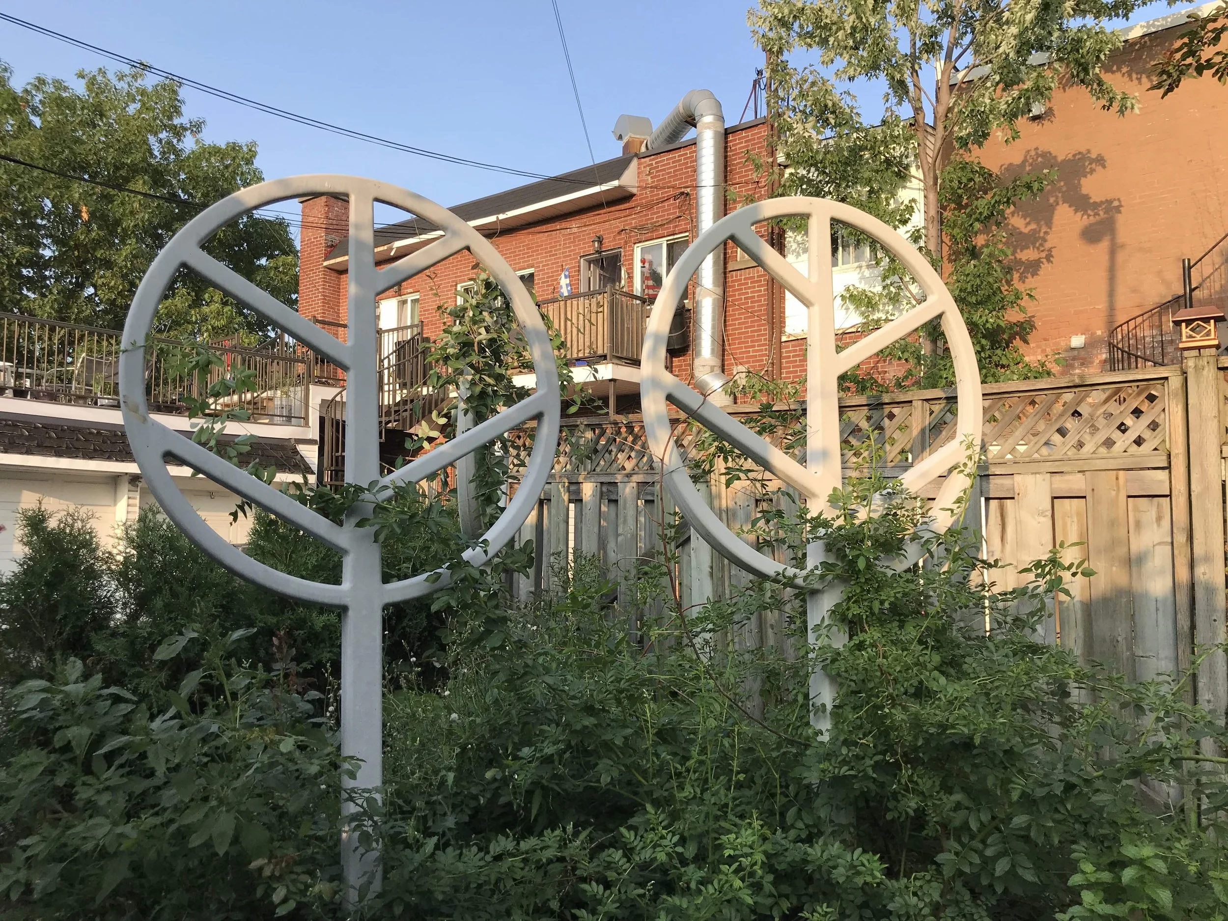 Deux grands cercles blancs en métal avec des lignes croisées, entourés de végétation, dans un jardin arboré, avec une maison en arrière-plan en briques rouges et une clôture en bois.