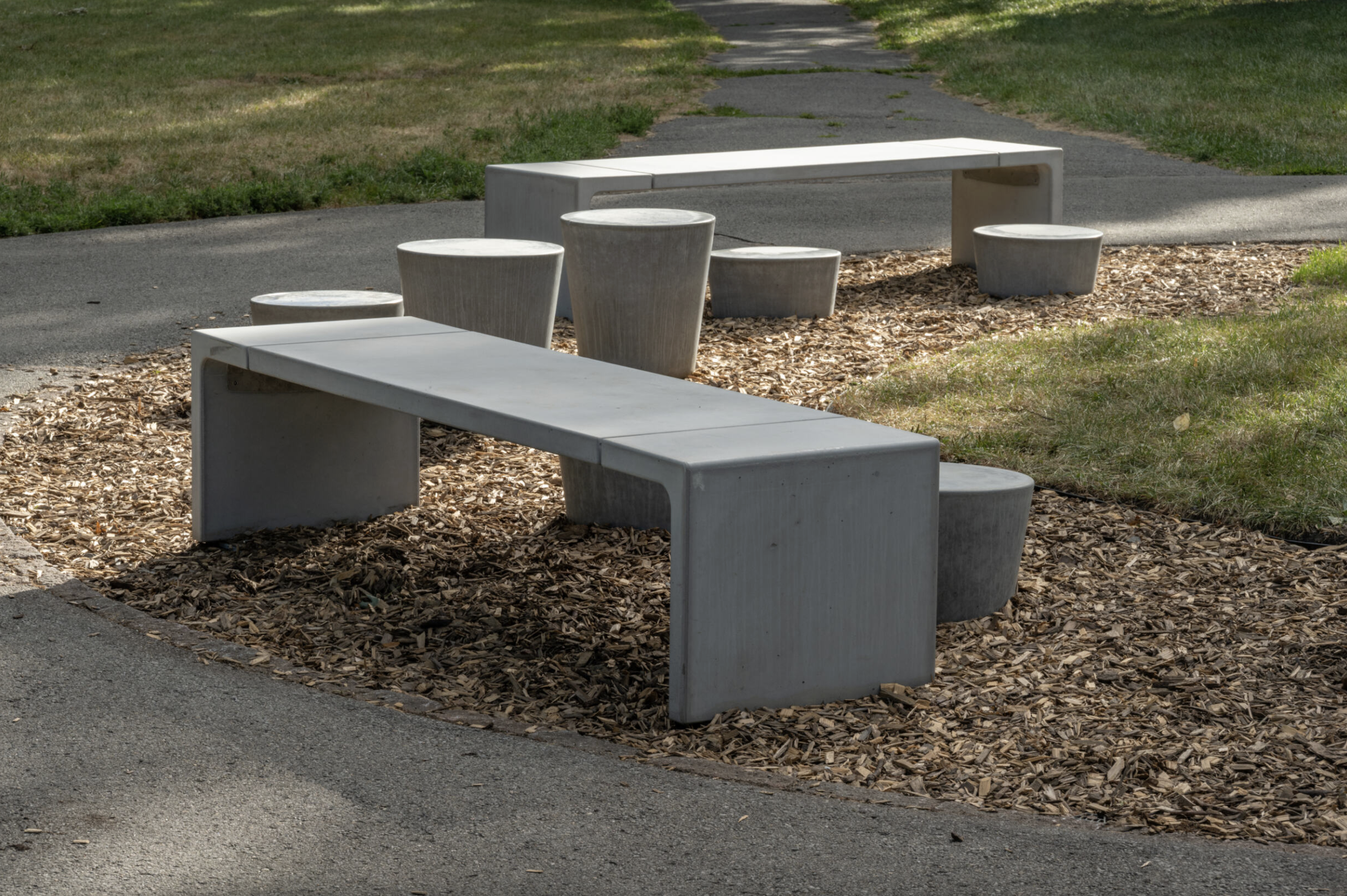 Bancs et tables en béton gris dans un parc, entourés de paillis de bois et d'herbe verte, avec un chemin en arrière-plan.