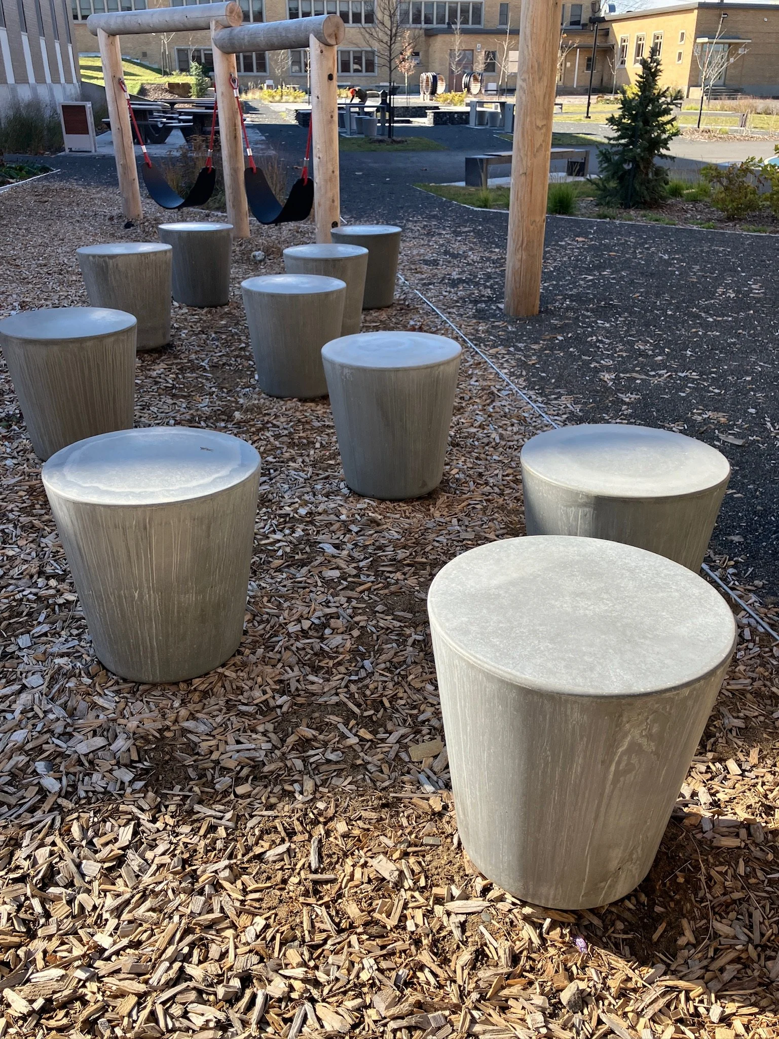 Mobilier de parc avec des tabourets en béton cylindriques, une aire de jeux avec deux balançoires en bois, et des bâtiments résidentiels en arrière-plan.