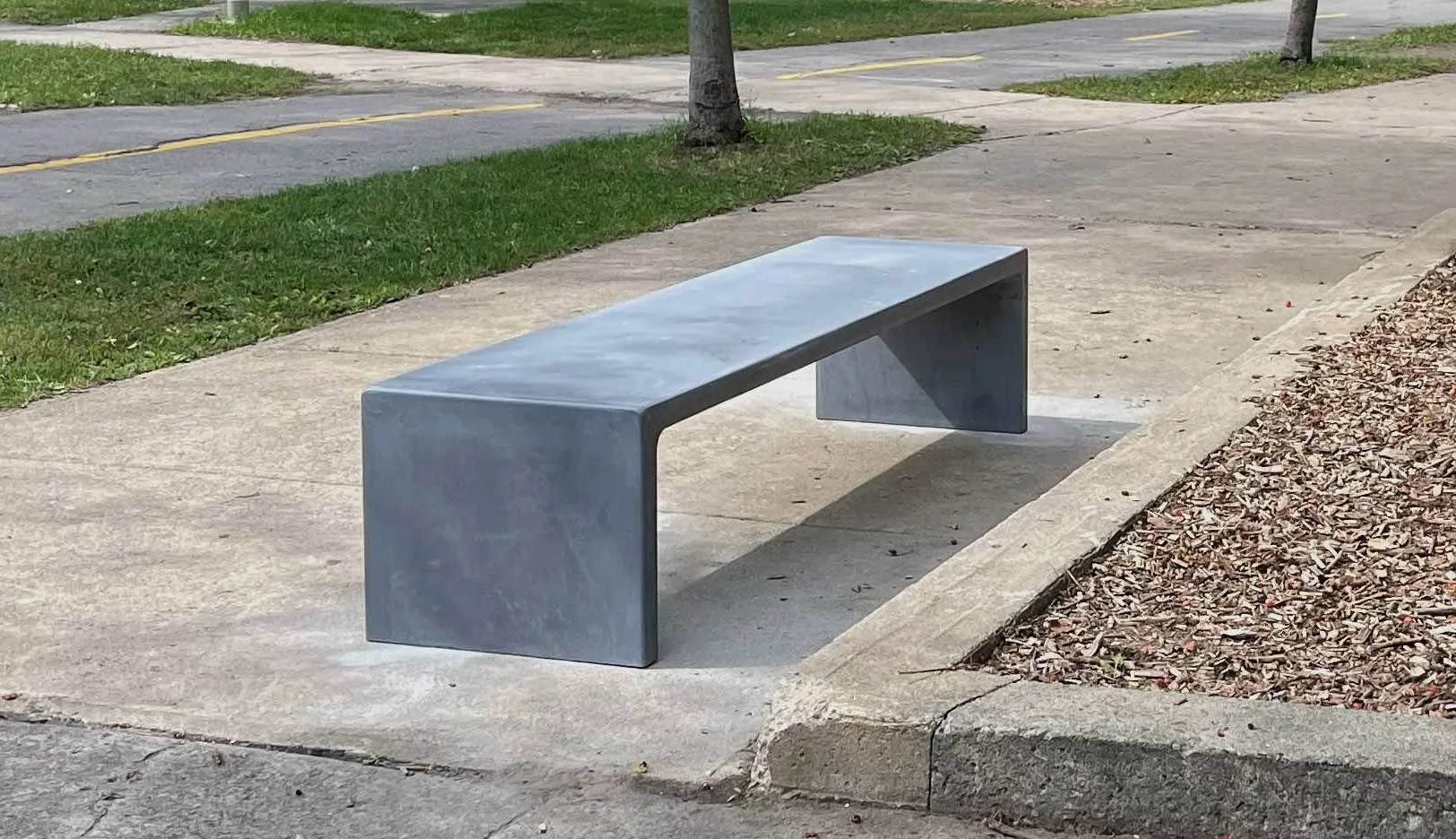 Banc en béton dans un parc, avec arbre et sentier en arrière-plan.
