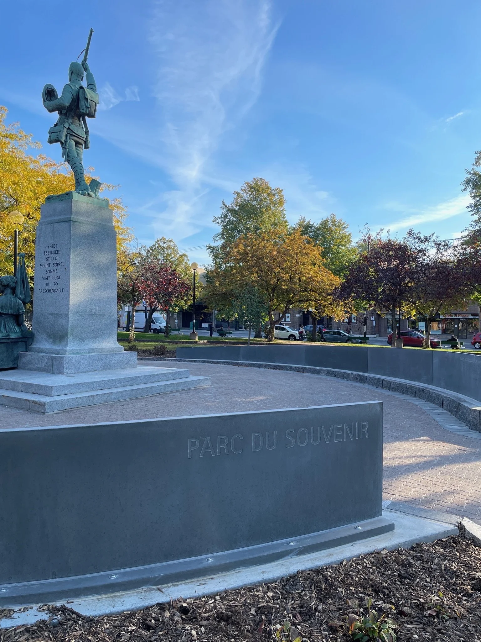 Un monument commémoratif dans un parc avec des statues, arbres et un ciel bleu. La plaque indique "Parc du Souvenir".