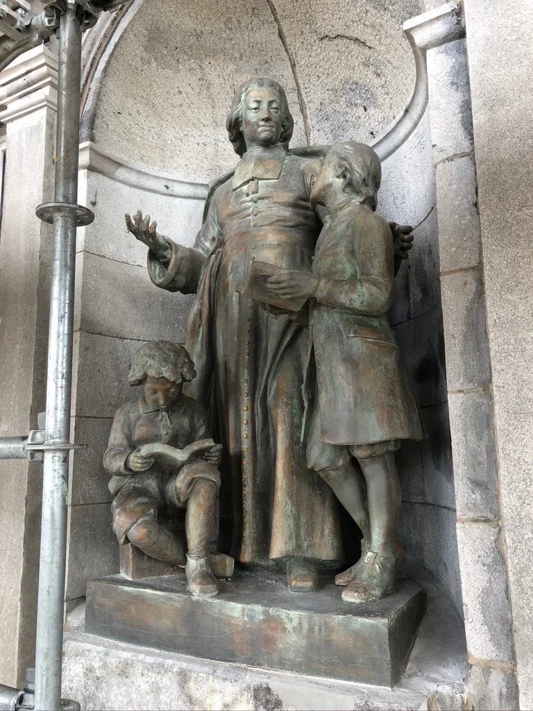 Statue en bronze représentant une femme avec deux enfants, dont un qui lit un livre et un autre qui regarde vers la femme, située dans un cadre architectural en pierre.