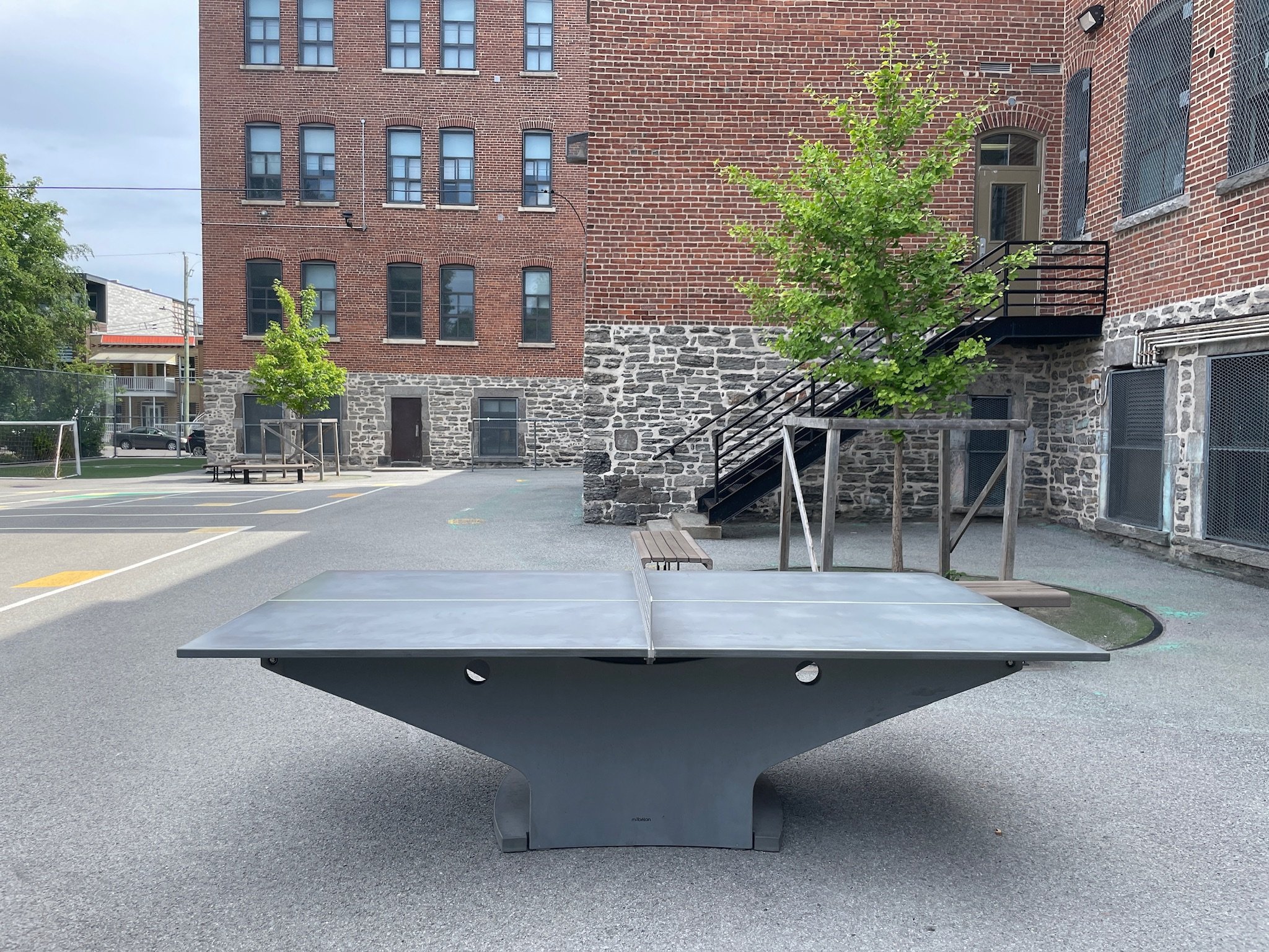 Table de ping-pong dans une cour d'école avec des bâtiments en briques en arrière-plan et deux jeunes arbres