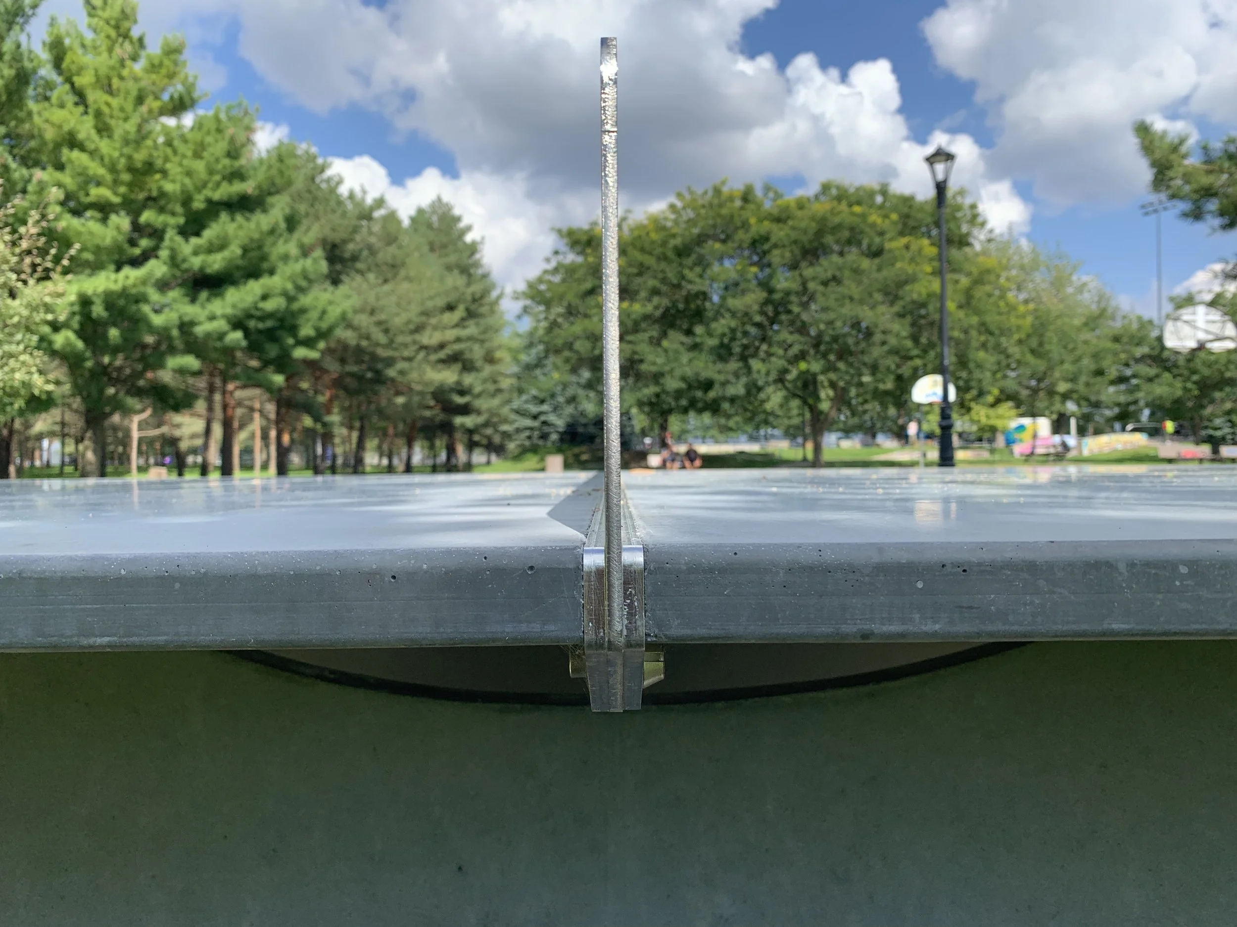 Table de ping-pong en extérieur avec vue sur un parc, arbres verts, lampadaire et bancs, ciel bleu avec quelques nuages.
