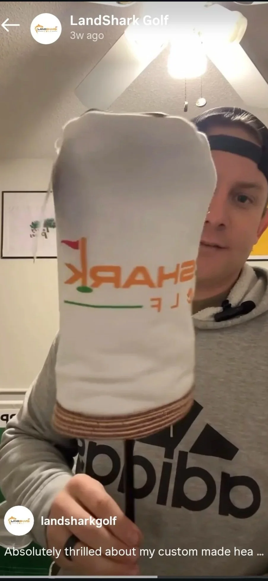 A man holding a Sharktale brew can toward the camera, with a ceiling fan overhead and a framed picture on the wall behind him.