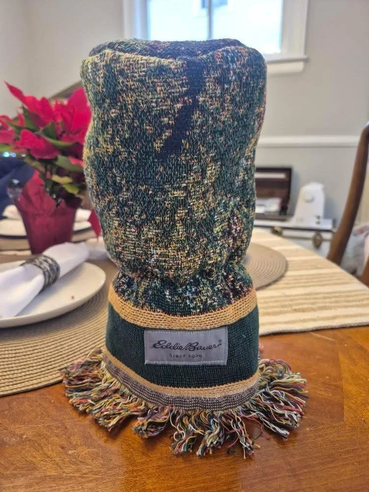 A colorful knit sock in dark green with multicolored specks, featuring a label that reads "LelliBauer Since 1928," placed on a wooden table with a poinsettia plant, a white plate with a napkin, and a window in the background.