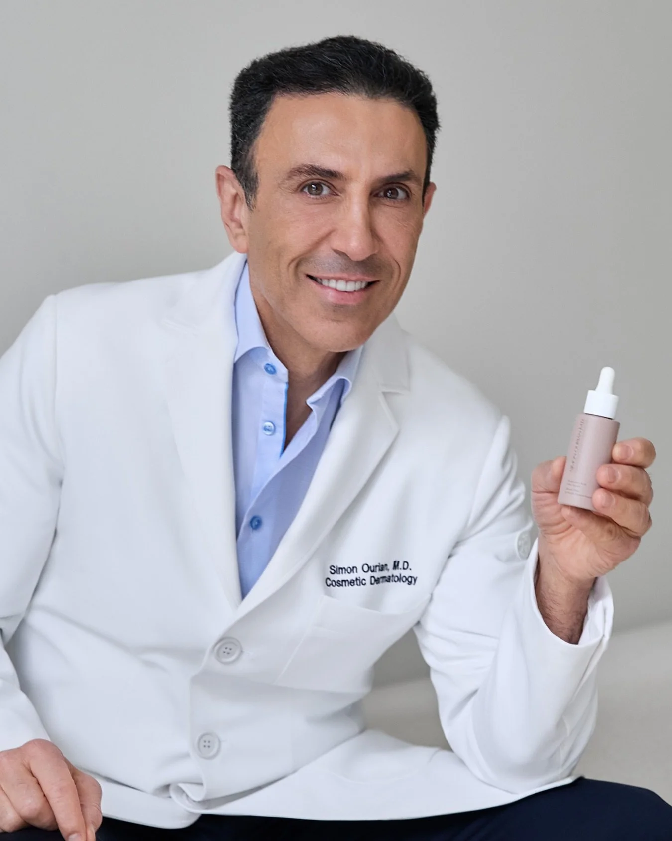 Un homme en blouse blanche, souriant, tenant une bouteille de soin cosmétique. La blouse a l'inscription 'Simon Ourian, M.D., Cosmetic Dermatology'.