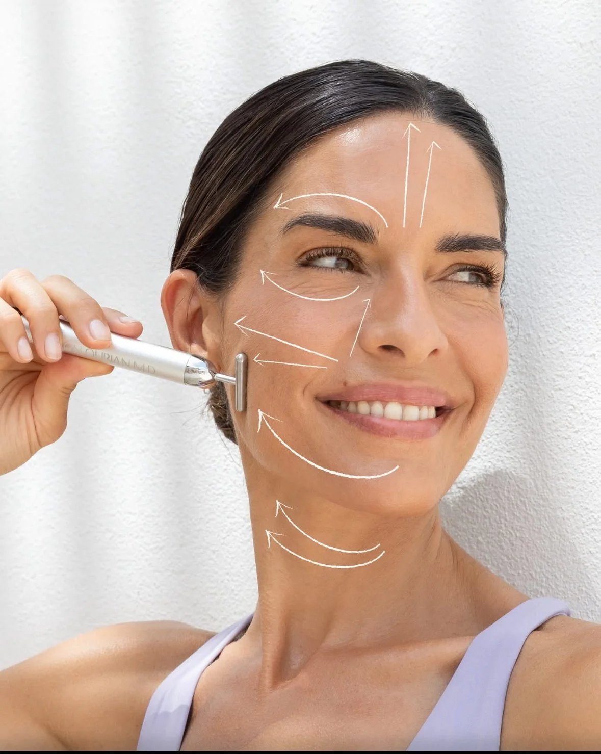 Une femme souriante effectuant un traitement de soin du visage à l'aide d'un appareil, avec des flèches blanches indiquant des zones de massage ou de traitement sur son visage, sa joue et son cou.