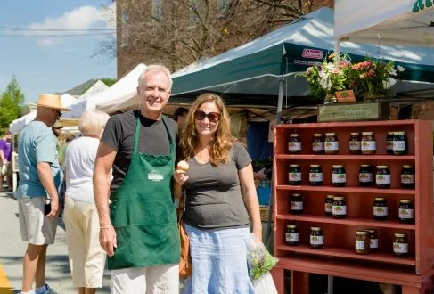 Two people, an older man and a woman, smiling and standing in front of a market stall with jars of homemade jam. The man wears a green apron, and the woman holds a bunch of green vegetables. Other shoppers and market tents are in the background on a 