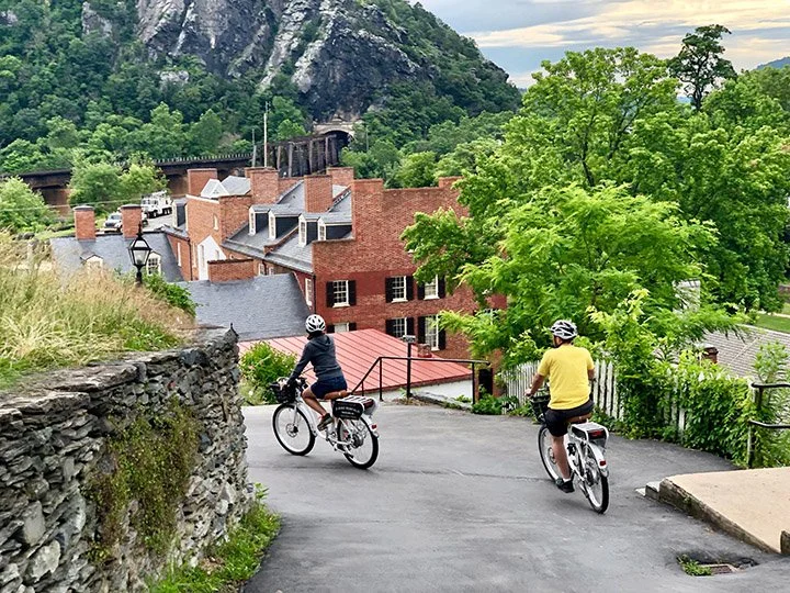 Two people riding bicycles uphill on a paved path, surrounded by greenery and old brick buildings with a mountain in the background.