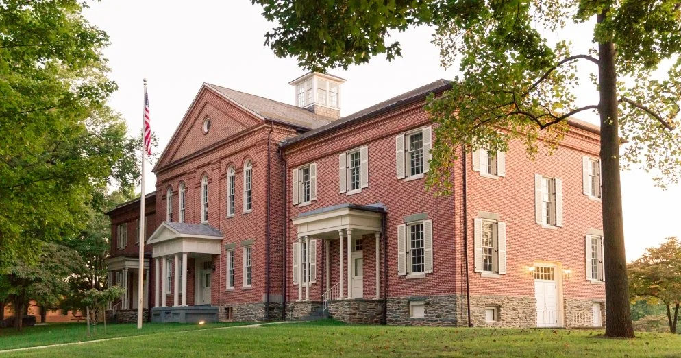 A large historic brick building with white shutters, a flagpole with the American flag, and surrounded by green trees and grass.