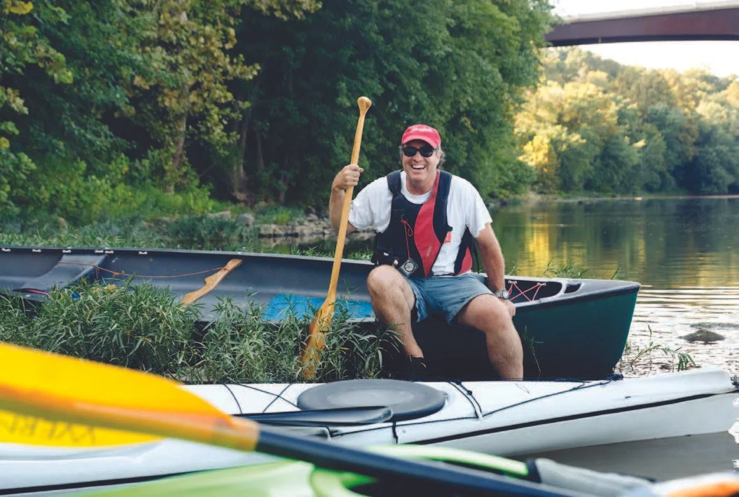 A man wearing a red cap and sunglasses sitting in a canoe on a river, smiling, holding a wooden paddle, with lush green trees in the background.