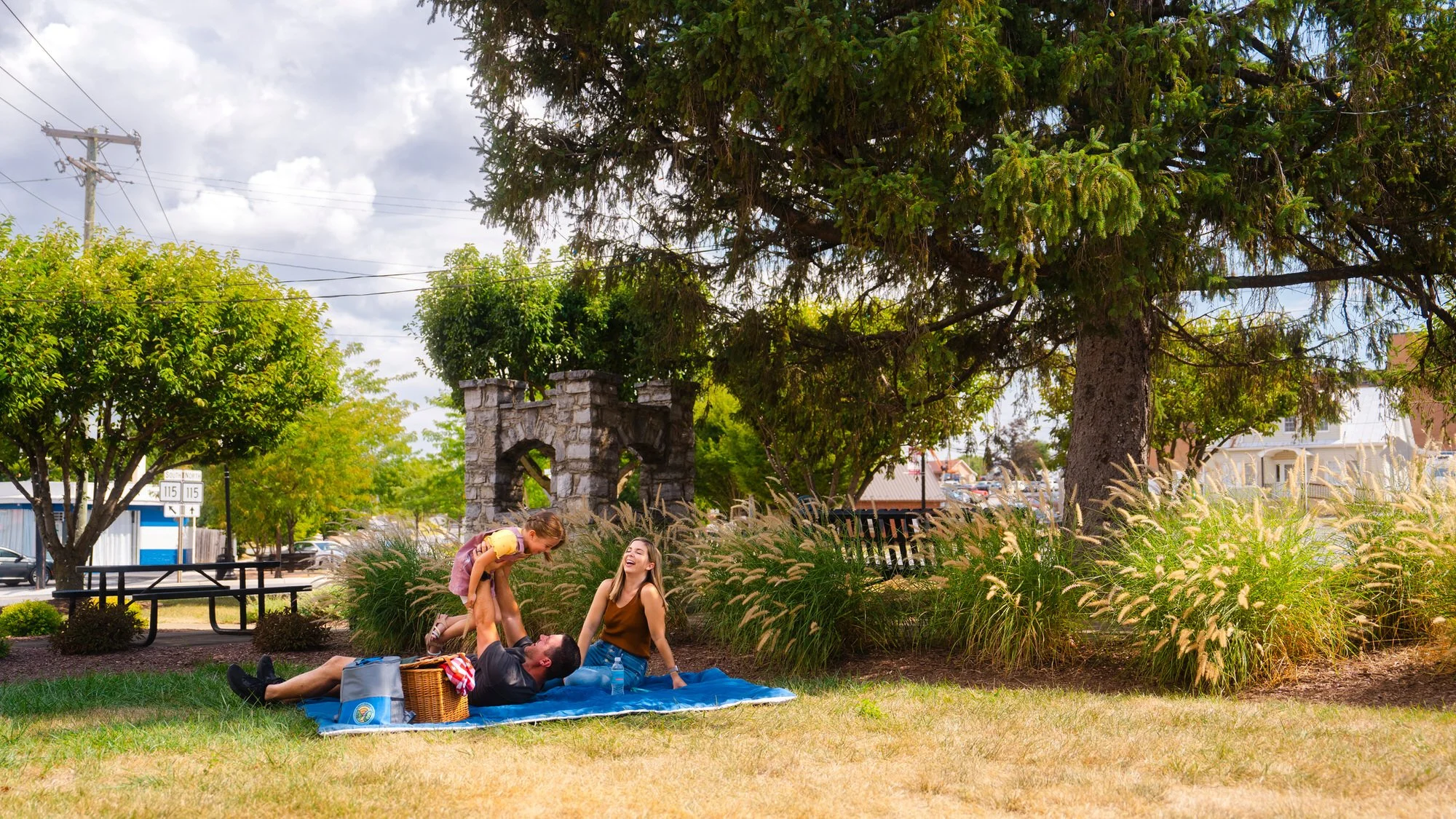 A family enjoying a picnic on a blue blanket in a park with trees, plants, and a stone archway in the background, under partly cloudy skies.