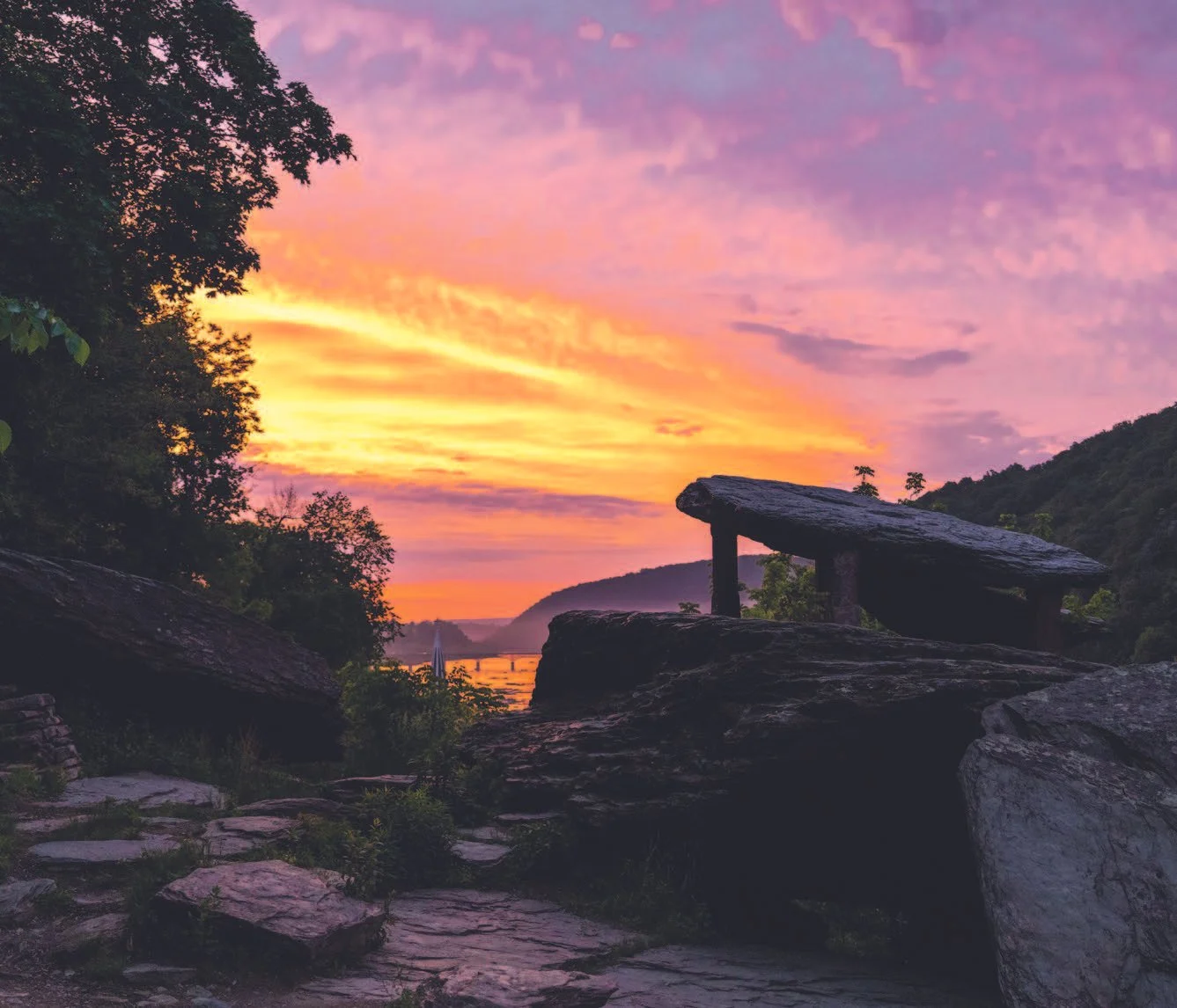 A sunset view over a river with a rock formation and a bench in the foreground, surrounded by trees and hills in the distance.