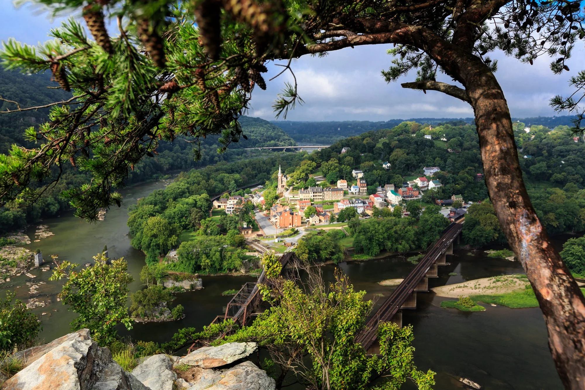 View of a small town nestled in a lush green valley with a river running through it, seen from a hillside partly obscured by tree branches.