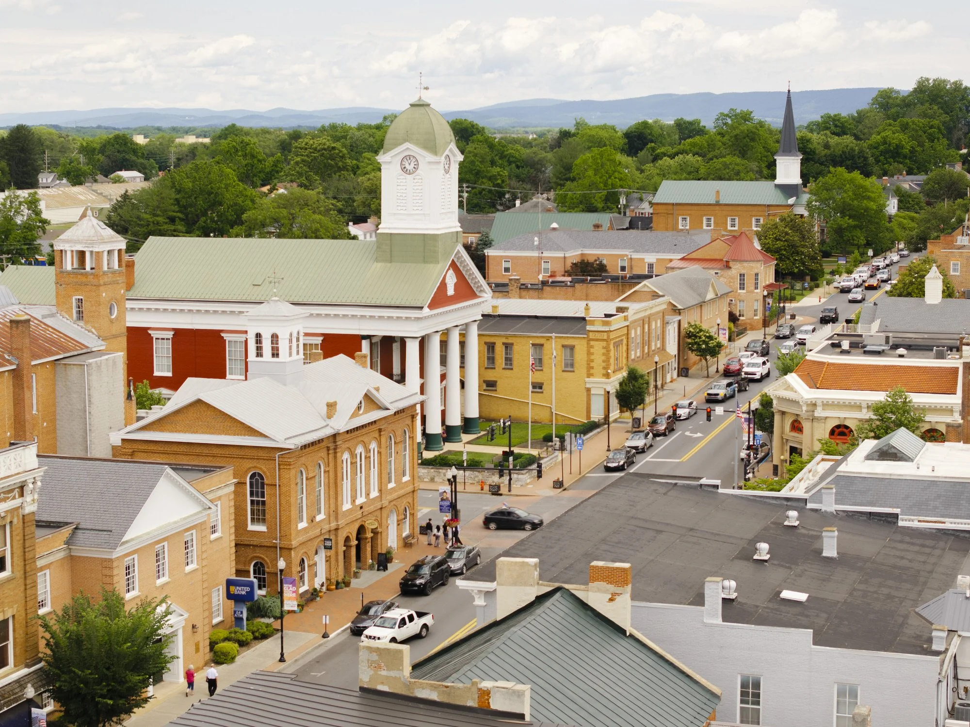 Aerial view of a small town with historic brick buildings, a church with a tall steeple, a courthouse with white columns, and tree-lined streets with parked cars, hills in the background, and a cloudy sky.