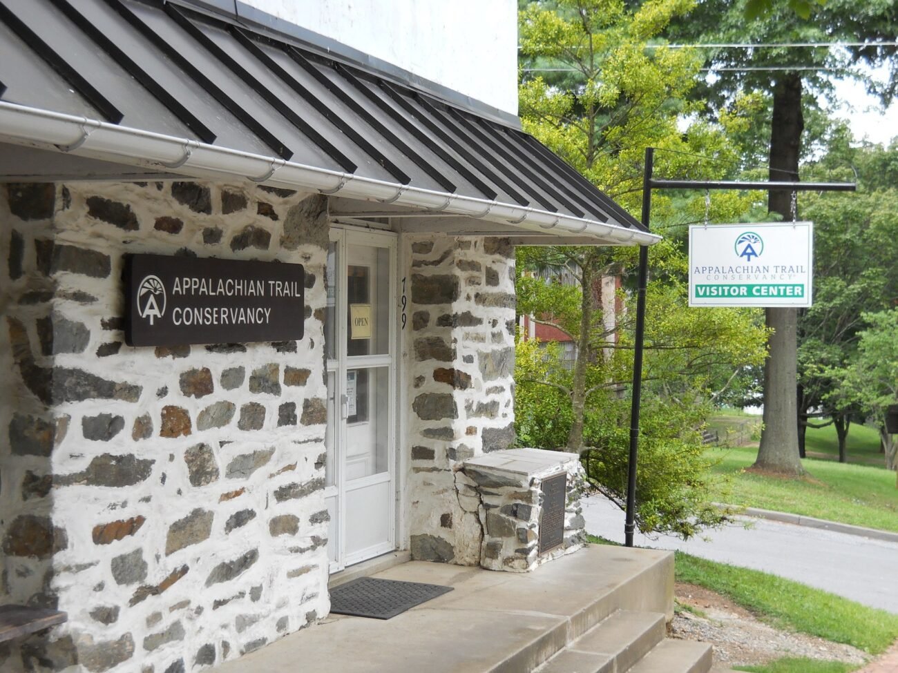 Stone building with 'Appalachian Trail Conservancy Visitor Center' sign, two steps leading to door, signboard outside, surrounded by trees.