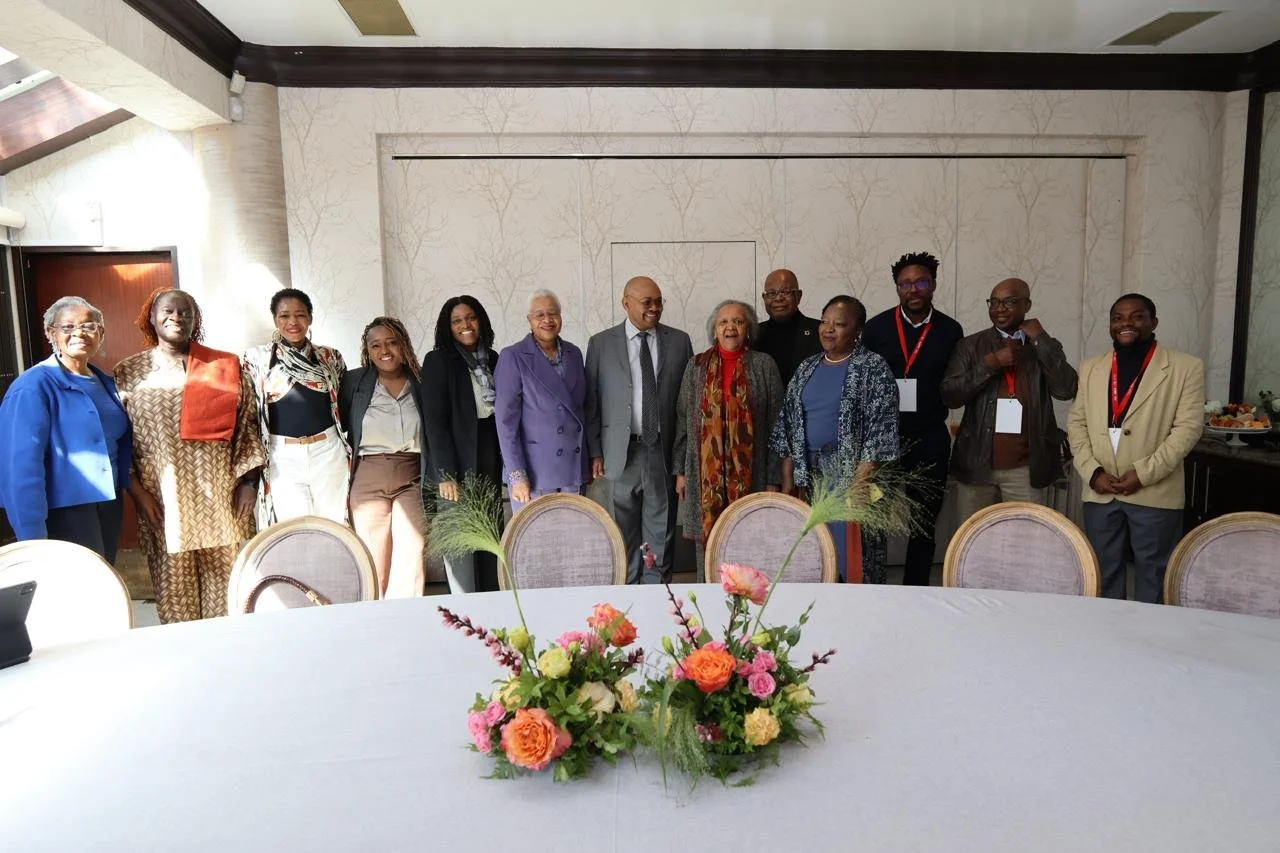 Group of diverse people standing in a line inside a well-lit room, with a floral centerpiece on a table in the foreground.