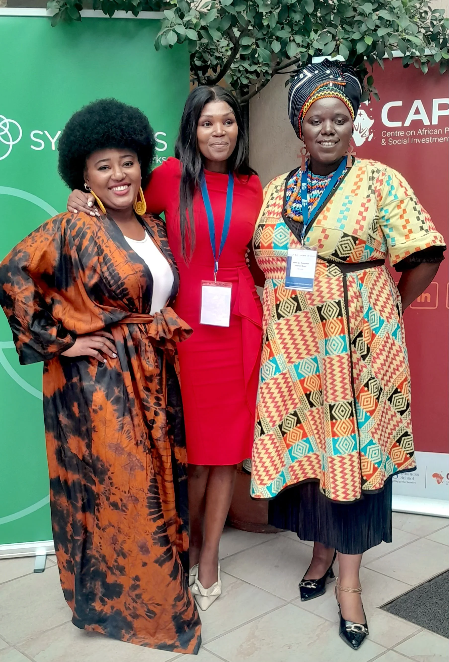 Three women standing together, smiling, at an event with banners in the background. One woman is wearing a colorful African print dress and a headwrap, the second woman is in a bright red dress, and the third woman is in a patterned dress with orange and black tones.