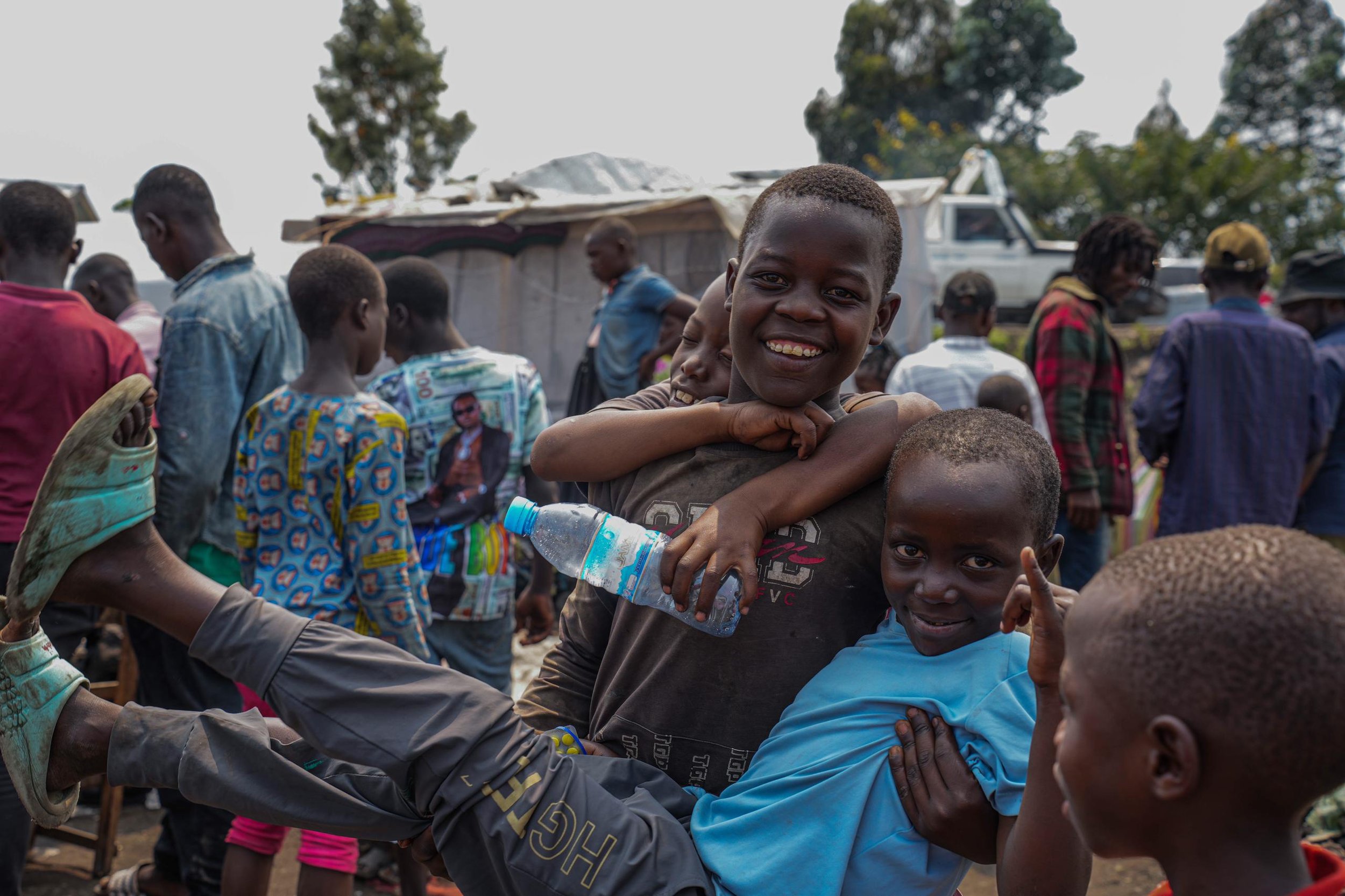 Smiling boy being carried by another boy amidst a crowd of children and adults outdoors.