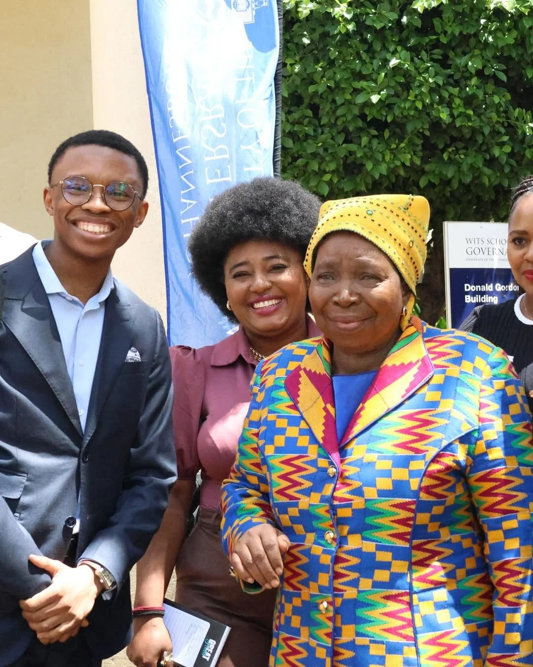 Four people standing outdoors, smiling, with a blue and white banner and green foliage in the background. The group includes a young man in a suit, two women, one in a colorful patterned jacket and headscarf, and the other in a maroon blouse.