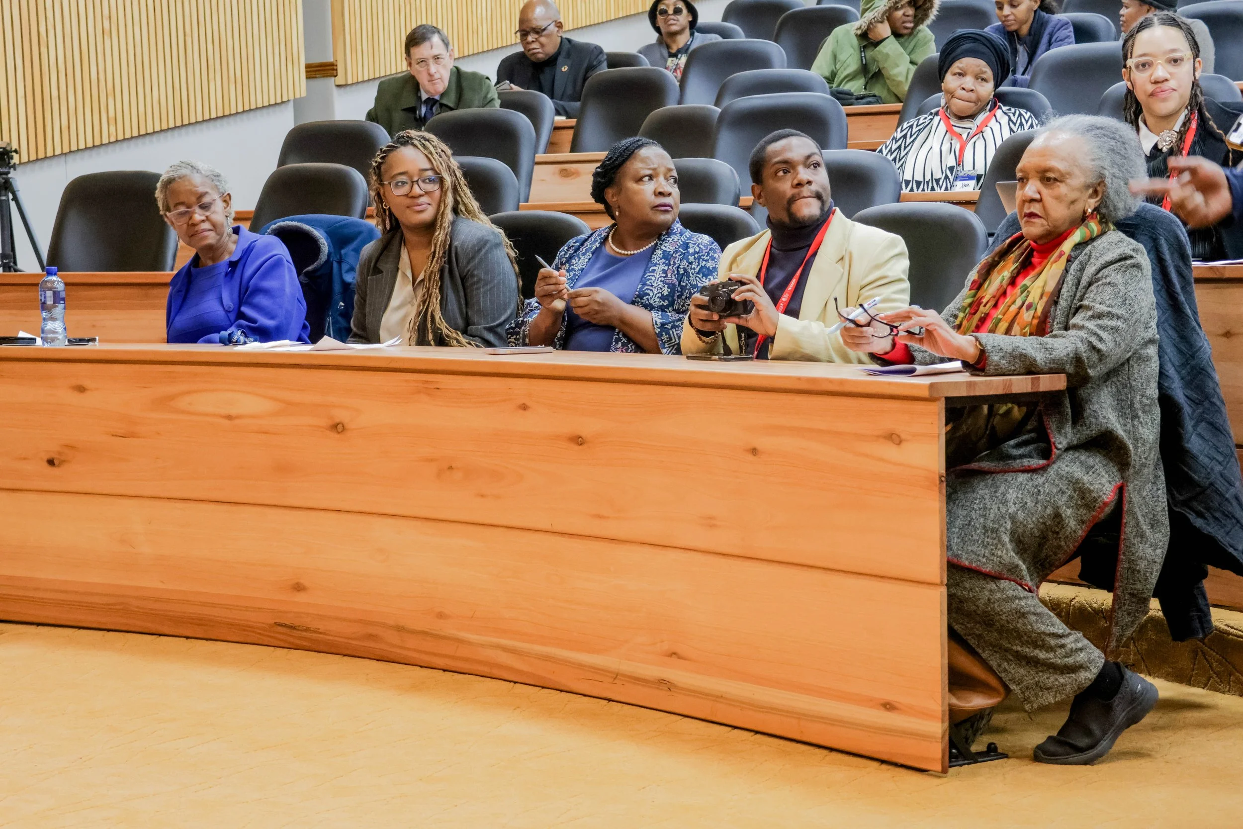 A diverse group of people seated in a conference room, some taking notes and others listening, with a wooden desk in the foreground.