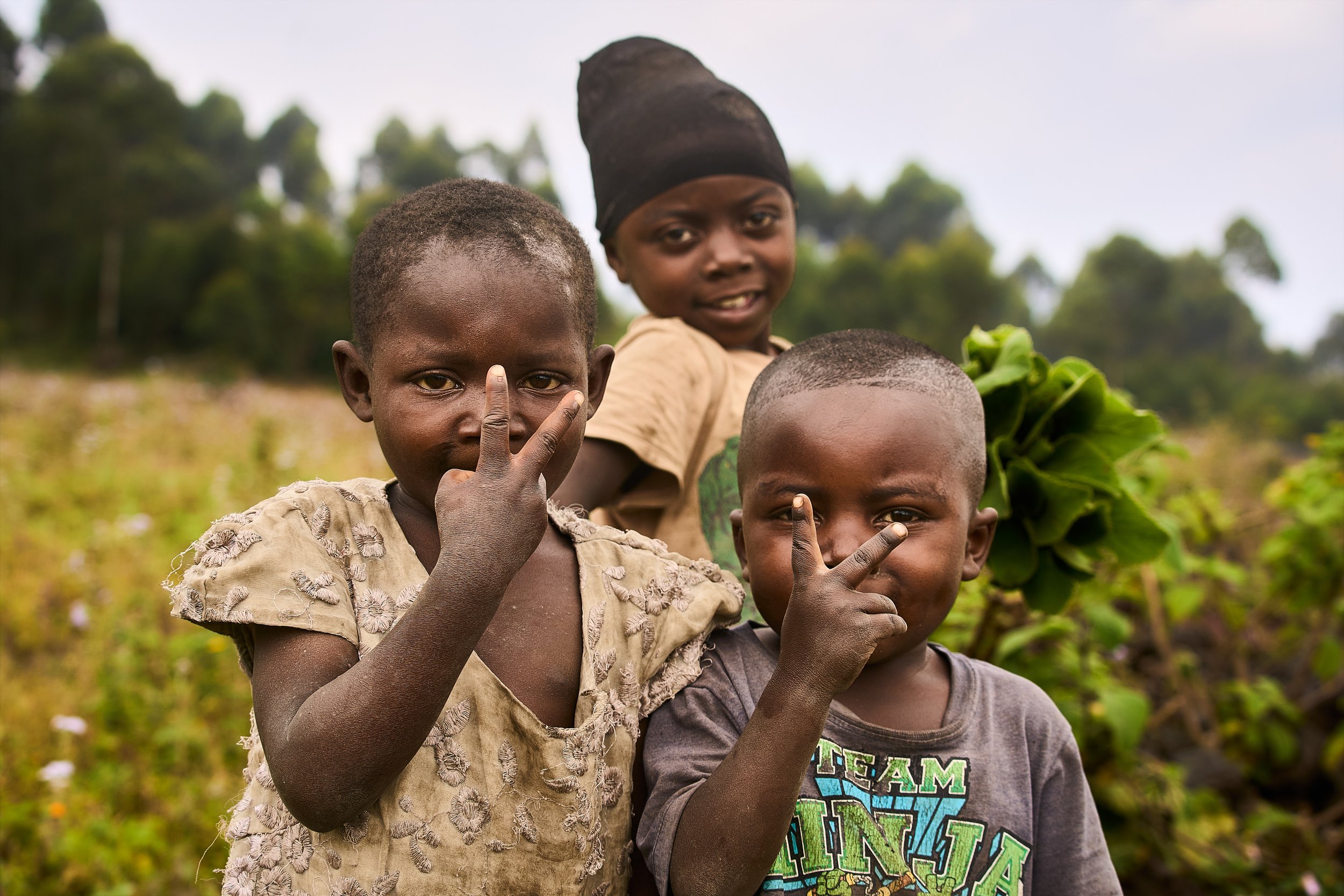 Three children in a field, two making peace signs with their hands near their faces, and one holding leafy greens, with trees in the background.