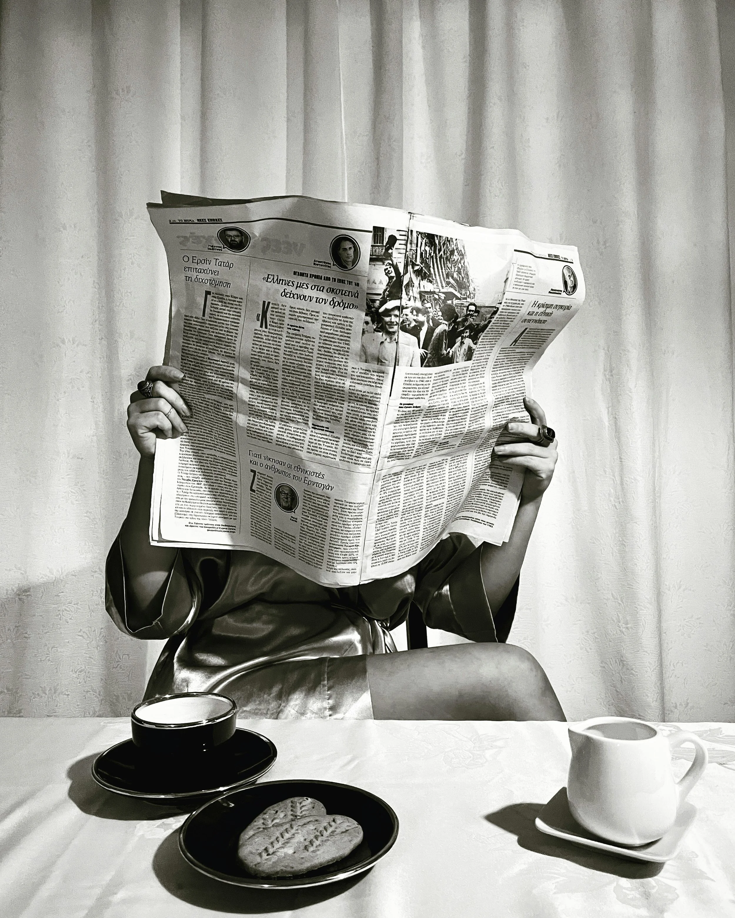 Person sitting at a table, reading a newspaper with their face obscured by the paper. On the table, there is a cup of coffee on a saucer, a plate with a cookie, and a small pitcher.