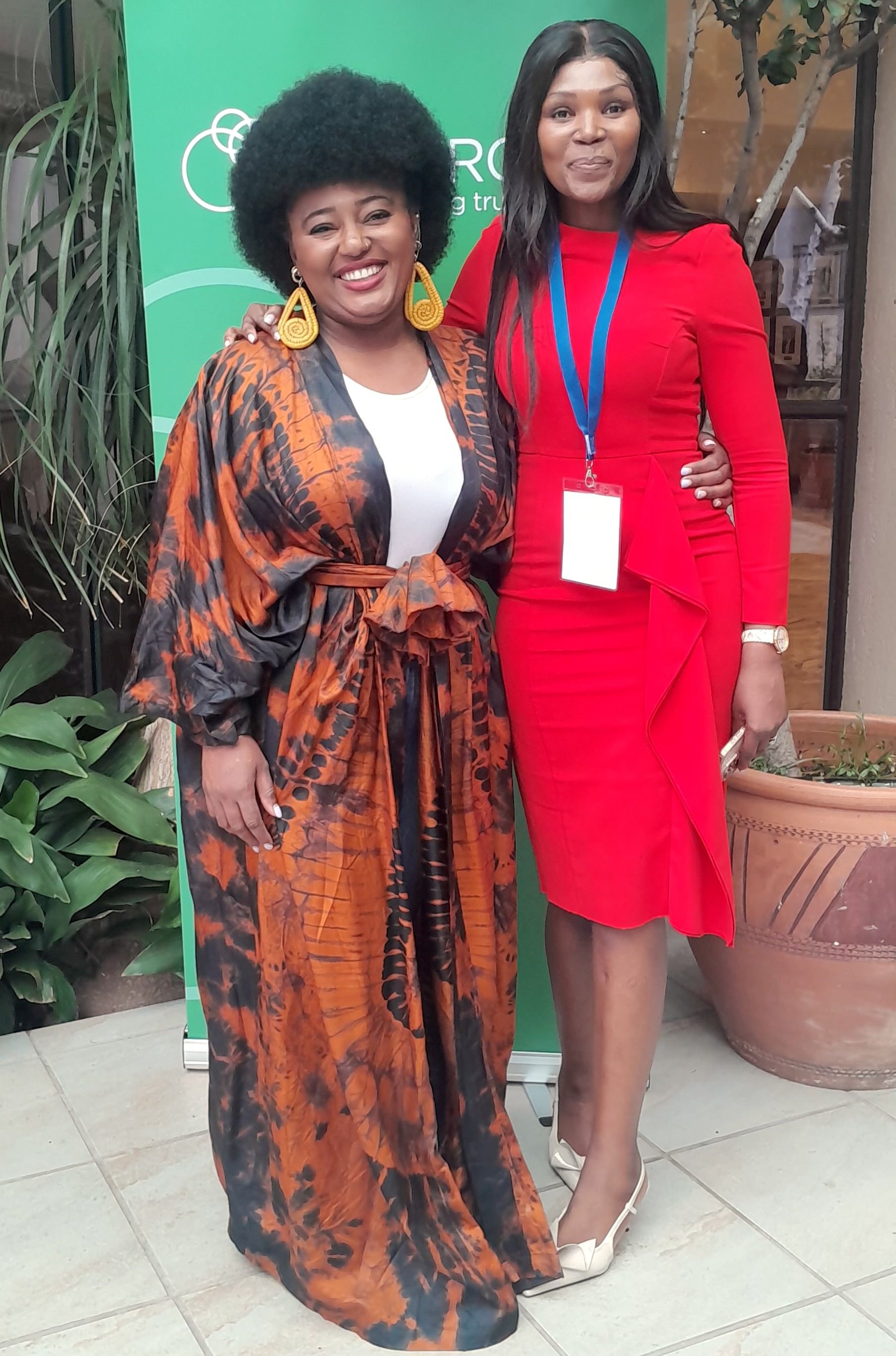 Two women standing together at an indoor event, smiling, with green background and plants around them. One woman is wearing a patterned orange and black dress with large earrings, and the other is wearing a red dress with a badge around her neck.