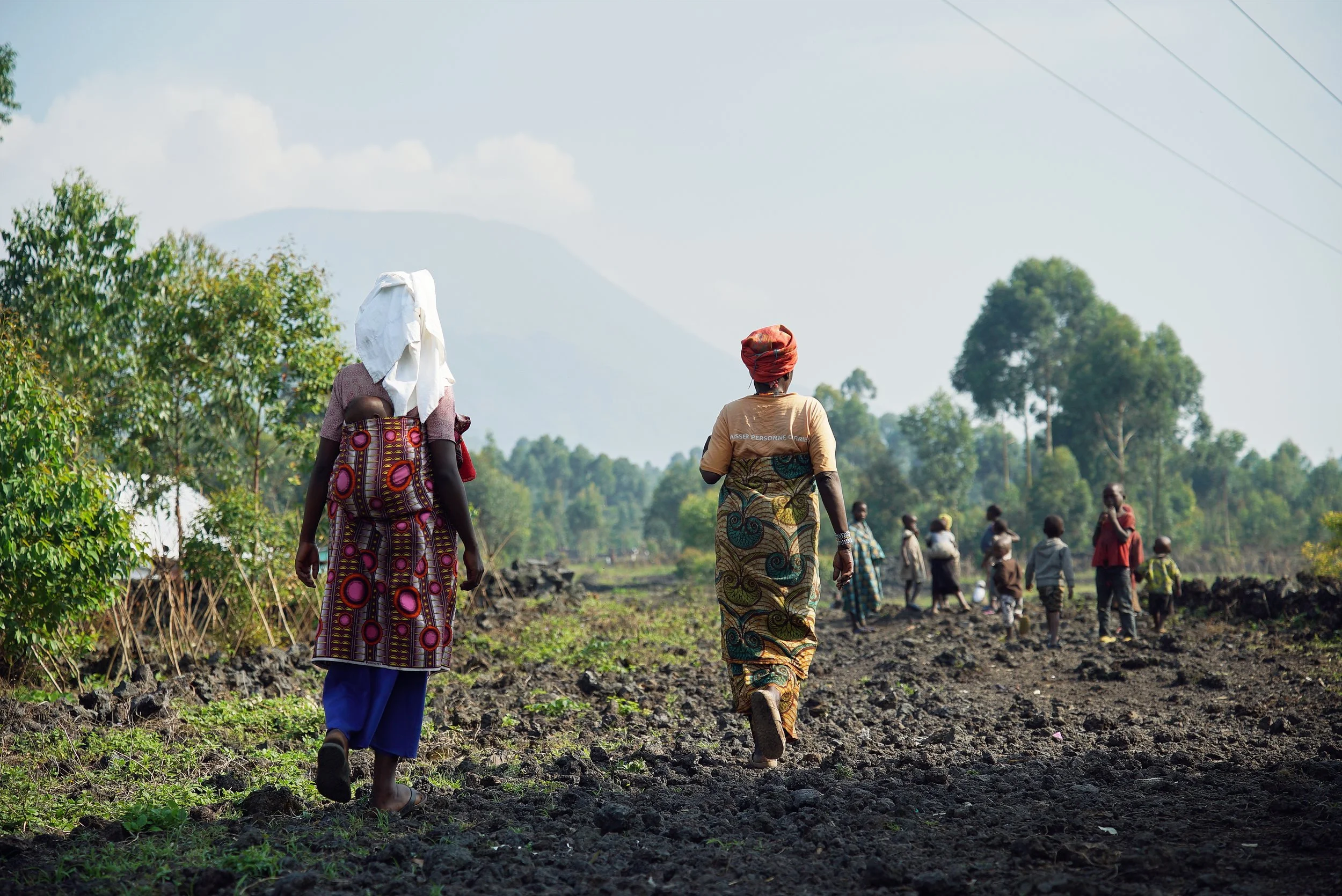 A group of people walking through a rural landscape with trees and mountains in the background, some wearing colorful traditional clothing and head coverings.