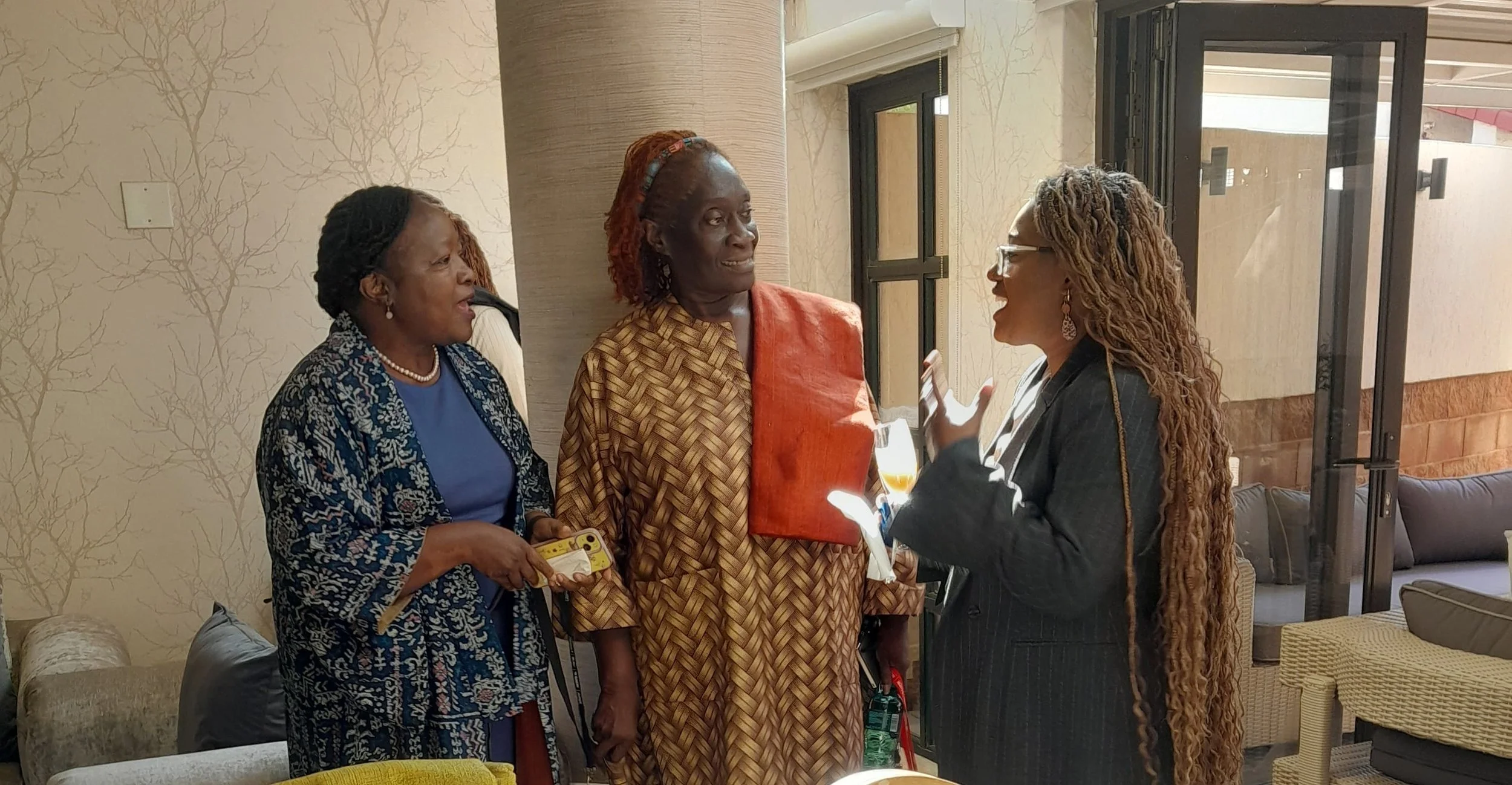 Three women engaged in a conversation indoors, with one woman gesturing with her hands and holding a wine glass, while the others listen. The background features a window and patterned wallpaper.