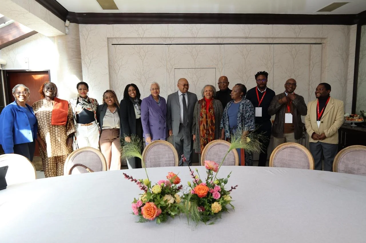 A group of 14 diverse people standing in a line behind a large table decorated with pink and orange flowers in a well-lit room with white walls and wood accents.